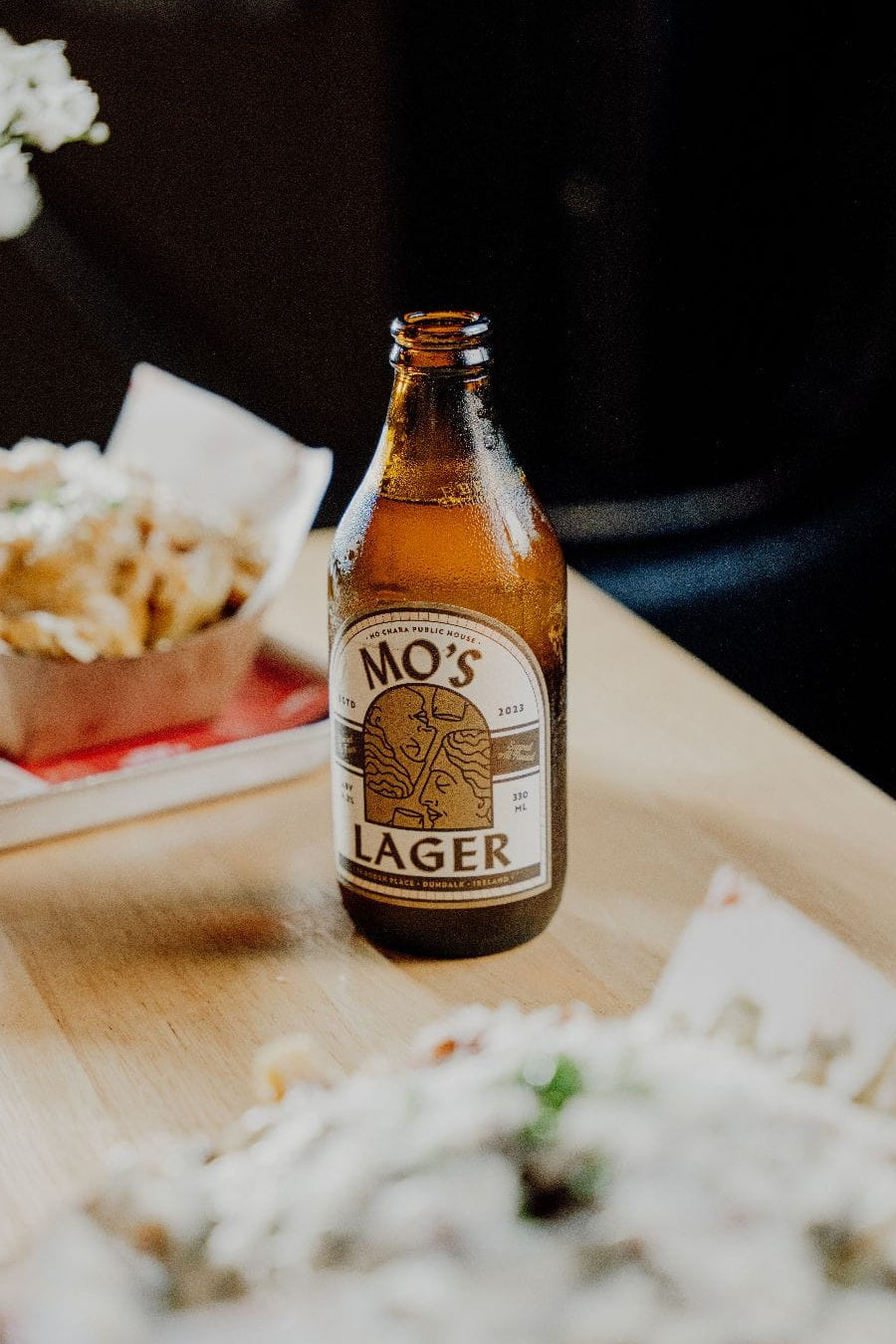 Bottle of Mo's Lager on a table with food and flowers in the background