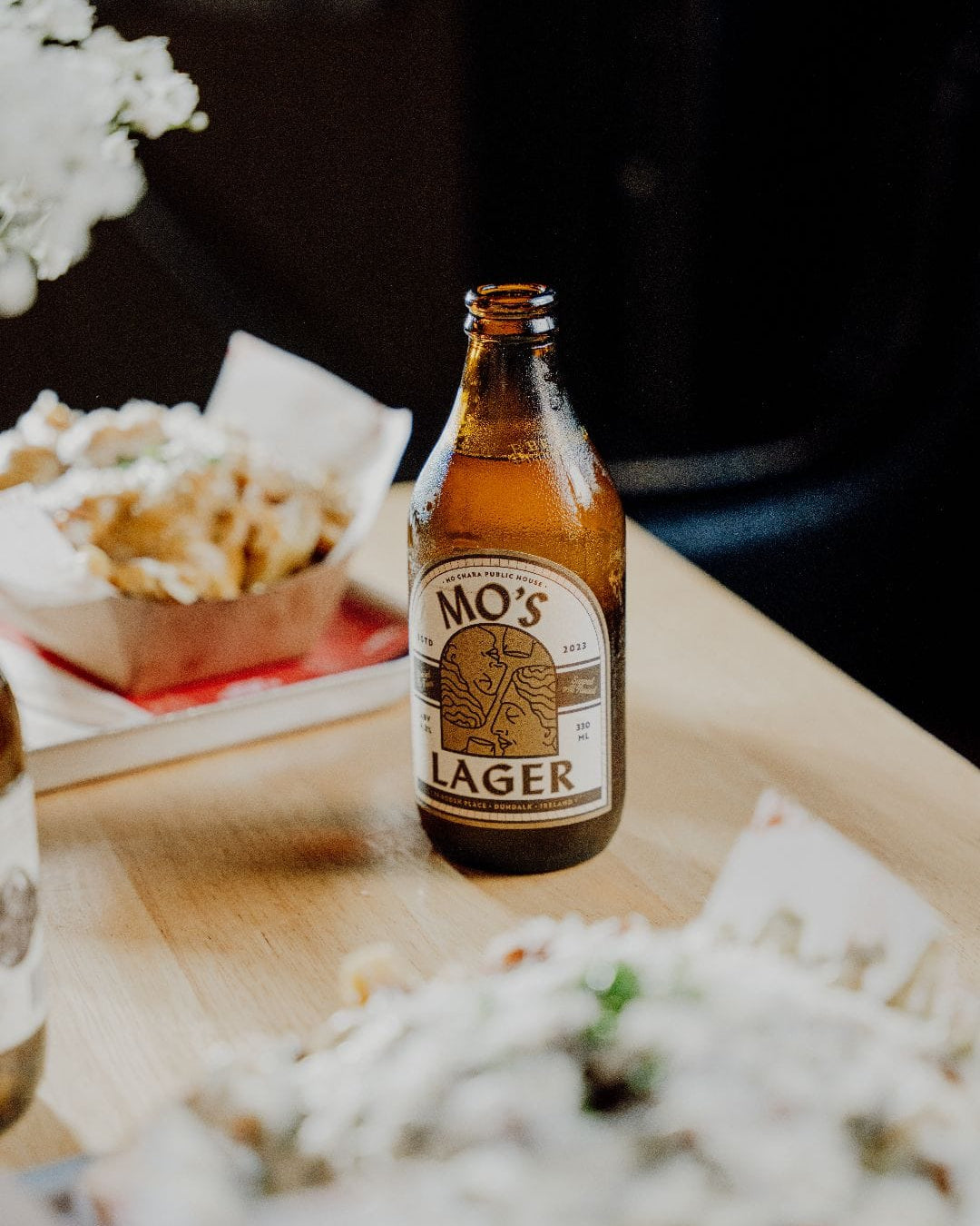 Bottle of Mo's Lager on a table with food and flowers in the background